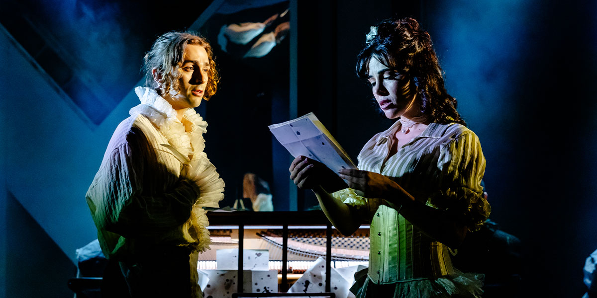 Two actors dressed in Victorian-era costumes perform on stage, depicting a scene where one actor is holding and reading a letter with an intense expression while the other looks on attentively.