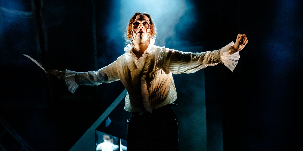 Person in a dramatic pose on stage, wearing a white shirt and vest, under dramatic lighting with a backdrop of mist.