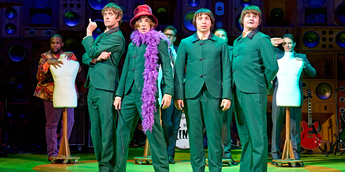 Four male performers in casual clothing are singing and playing instruments on stage, with bright lighting.