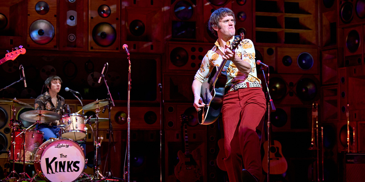 A man with short brown hair sits on a stool, playing a guitar, surrounded by warm, golden lighting.