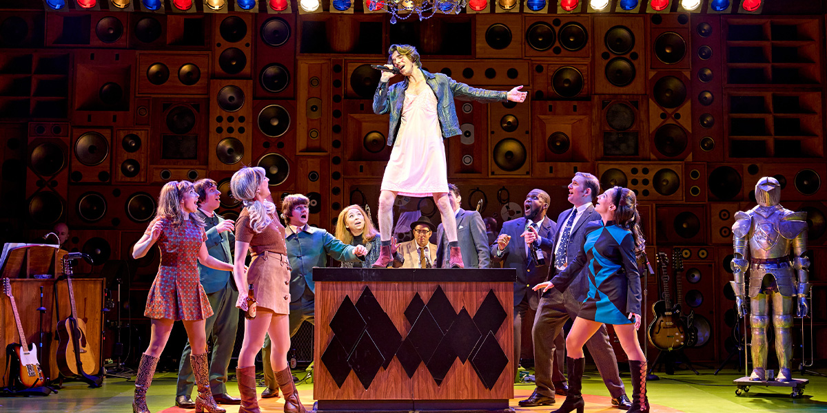 A group of musicians perform on stage, dressed in casual attire, with bright lighting and a vibrant backdrop.