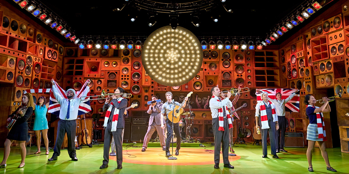 A group of performers in vibrant costumes poses joyfully on stage, with bright lighting and a colourful backdrop.