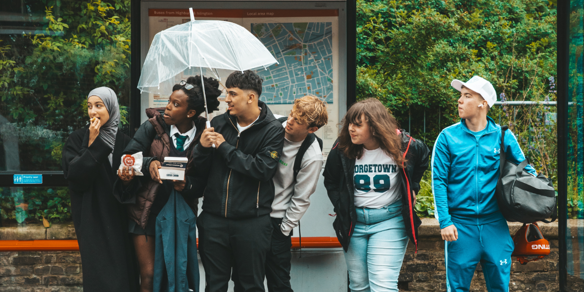 Six individuals are waiting at a bus stop under a light rain, with one of them holding an umbrella. They are standing in front of a bus route map.