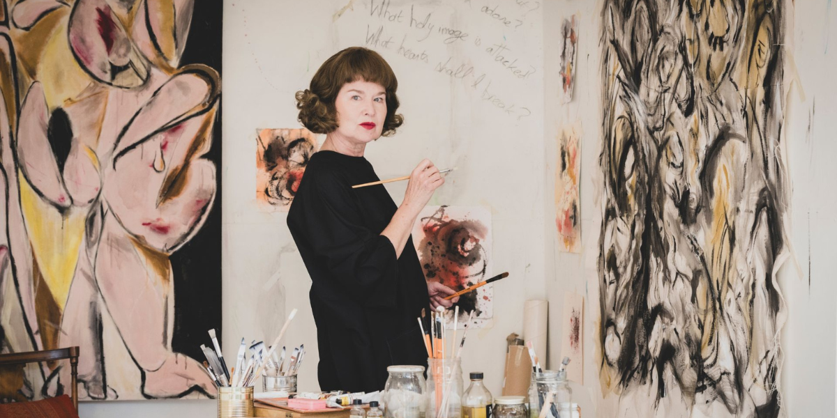 Artist standing in front of large canvases with abstract artwork, holding a paintbrush in a studio filled with painting supplies.