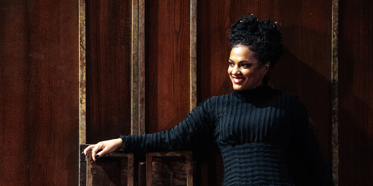 A scene from Twelfth Night featuring an actor in a black dress standing against a wooden panelled backdrop