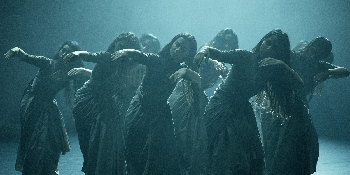 A group of dancers performs a synchronized routine on a foggy stage, dressed in flowing, dark attire.