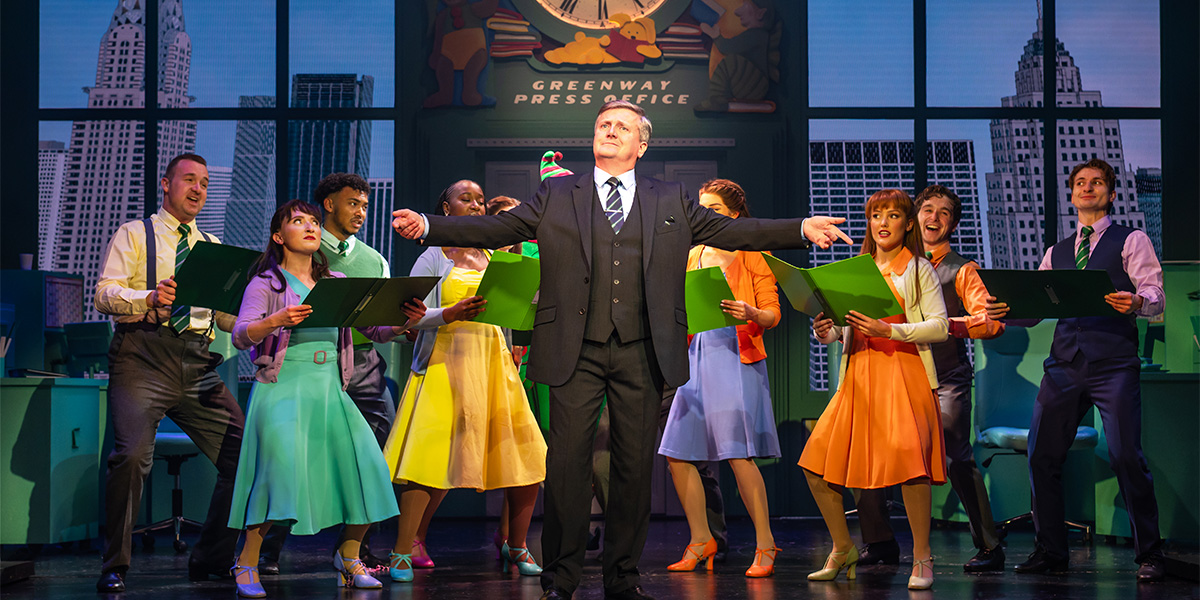 Aled Jones as Walter Hobbs stands centre stage with the cast of Elf the Musical, smiling.