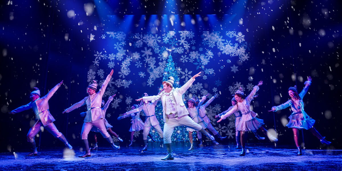 Joel Montague as Buddy stands centre stage with the cast of Elf the Musical, vibrant costumes visible.