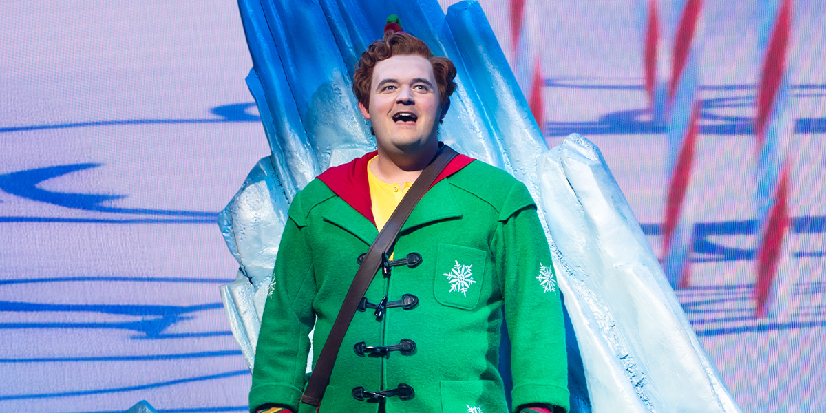 Joel Montague as Buddy in a green elf costume, smiling, with festive decorations in the background.