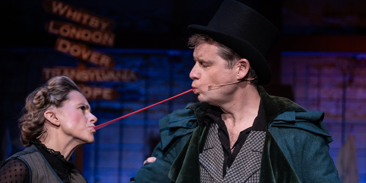 Two people stand opposite one another a piece of what looks like chewing gum stretches from one of their mouths to the other.