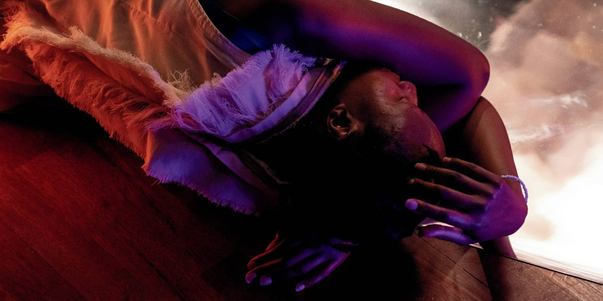 A person lying down with an arm bent over their eyes, dressed in a fluffy feathered outfit, under colourful lighting.