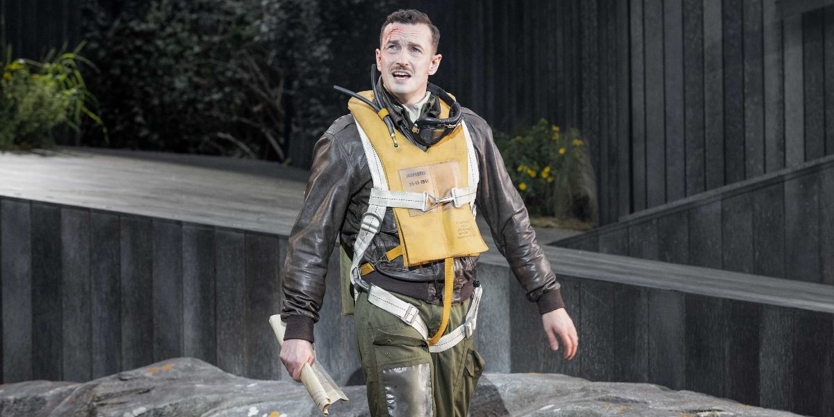 A man in a brown leather jacket and yellow life vest stands outdoors holding a rolled-up paper, with a serious expression. The background features dark, modern architecture and some greenery.