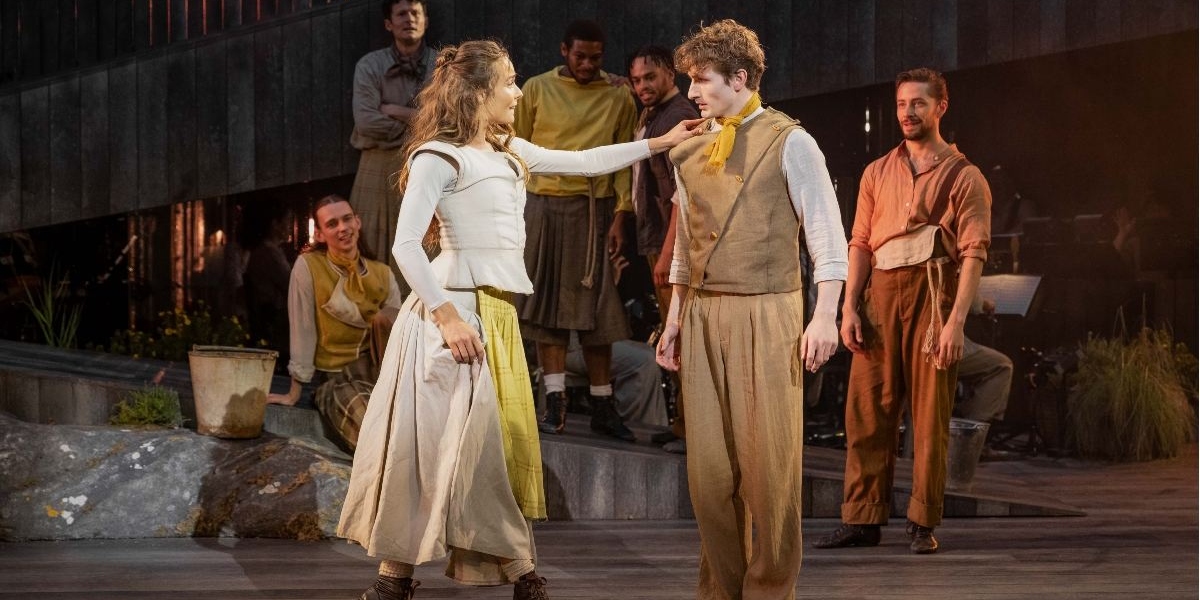 Two actors perform on stage in rustic costumes, with a woman touching a young mans chin. Several other actors watch in the background. The set has earthy tones and a naturalistic design, evoking an outdoor rural setting.
