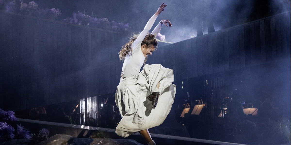 A woman in a long, flowing skirt and white top dances energetically on stage, one arm raised and one leg lifted. Dramatic lighting and mist create a theatrical atmosphere, with musicians visible in the background.