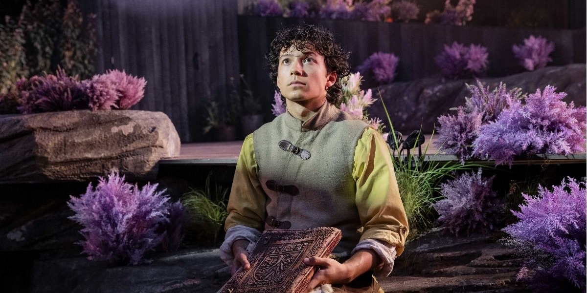 A young person sits among purple foliage and rocks, holding an ornate book and looking upwards with a thoughtful expression. The scene appears theatrical or fantasy-themed.