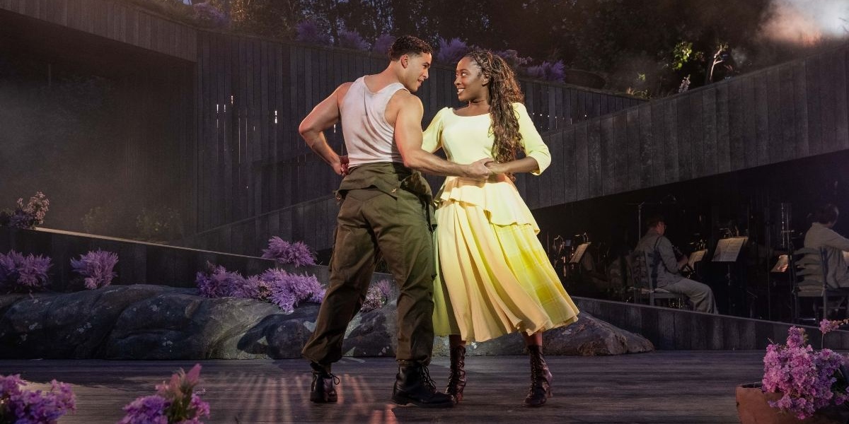 A man in a white tank top and green trousers dances with a woman in a flowing yellow dress on an outdoor stage decorated with purple flowers; musicians are visible in the background.