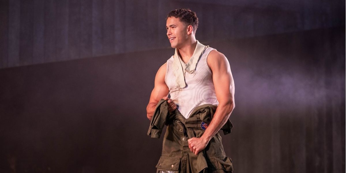 A muscular man in a white tank top and green trousers stands on stage, holding a green jacket and smiling confidently under dramatic lighting.