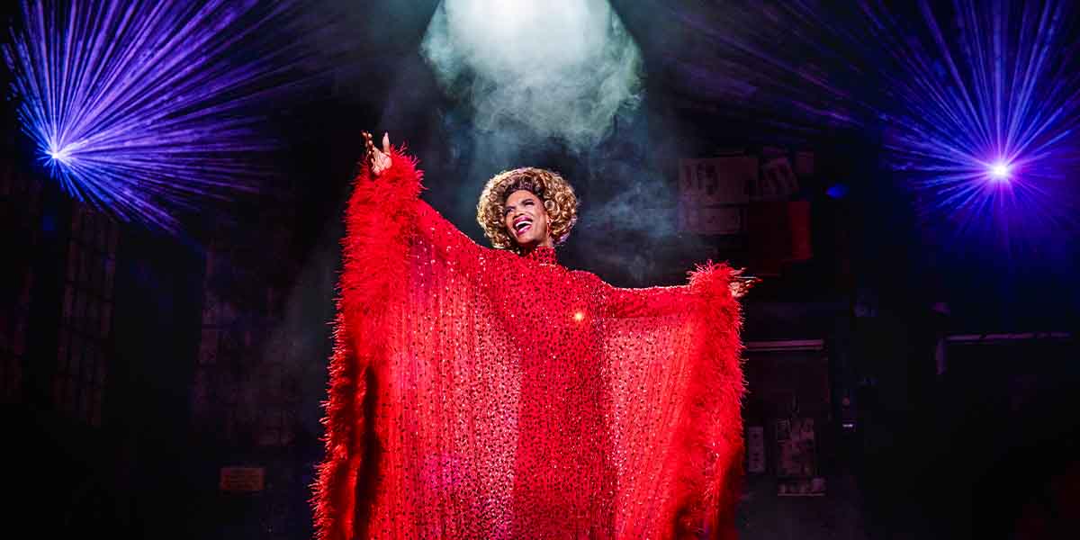 Johannes Radebe as Lola, wearing a sparkling red outfit, performs under dramatic lighting and smoke.