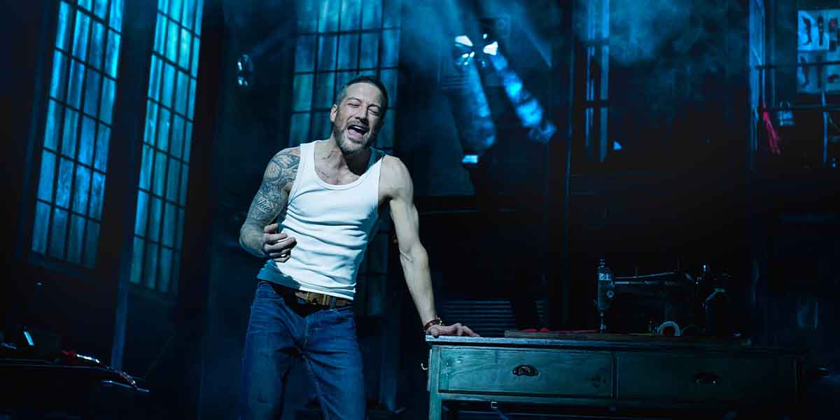 Matt Cardle as Charlie Price sings passionately in a dimly lit, industrial-themed stage setting.