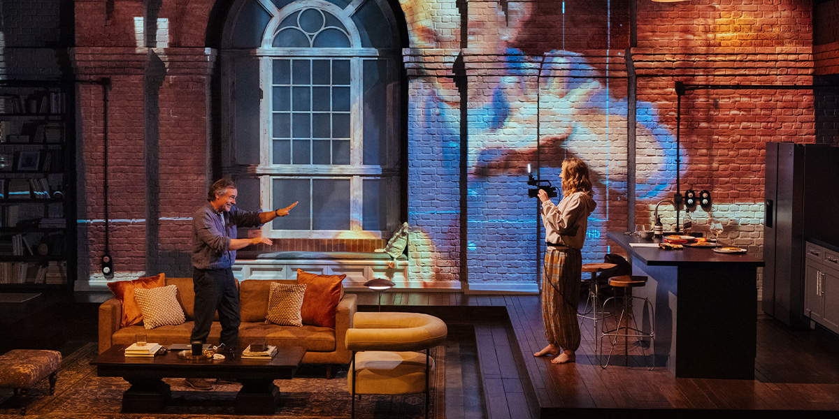 Two actors performing on stage in a set designed to resemble a cozy living room with brick walls and modern furnishings.