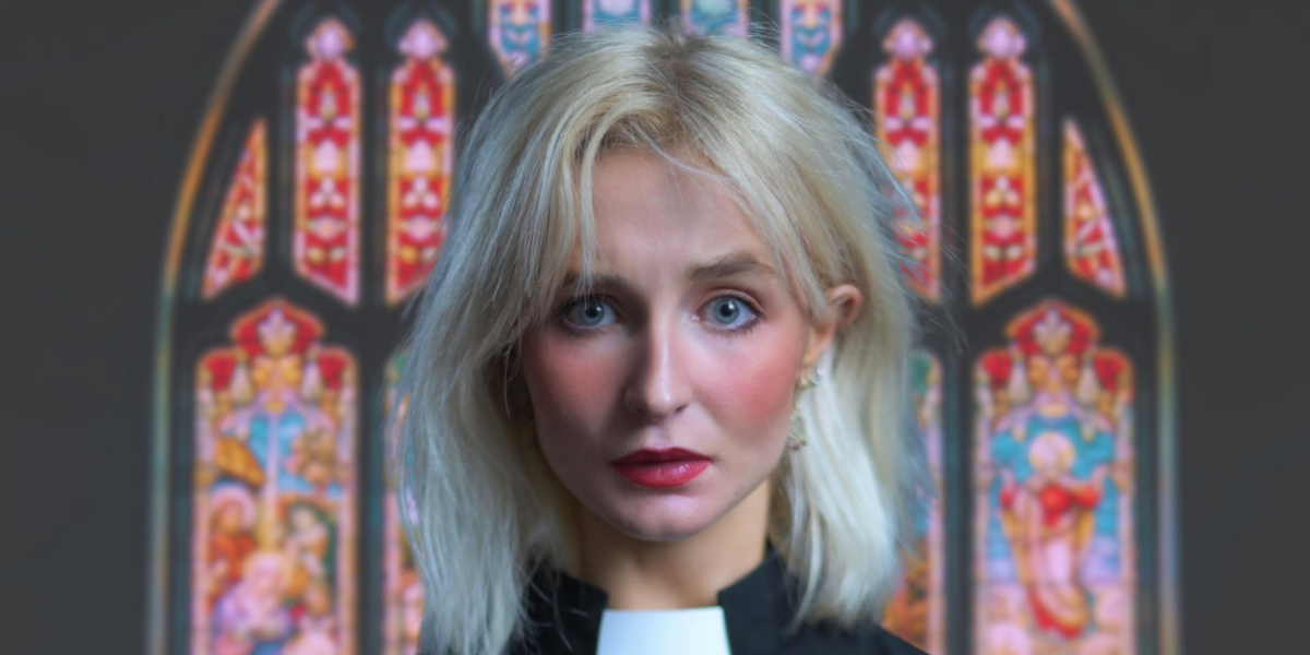 A woman with blonde hair and a serious expression, wearing a clerical collar, stands in front of a colourful stained glass church window.