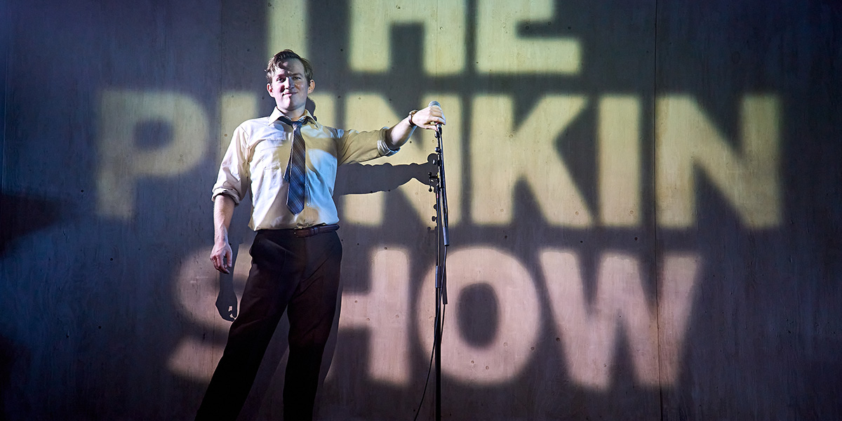 Jack Holden stands confidently in a dramatic pose, wearing a dark costume against a moody, dimly lit background.