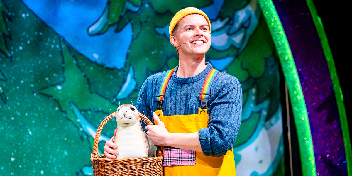 Matt Kennedy as Beast, wearing a blue coat, stands in a dimly lit castle interior, looking contemplative.