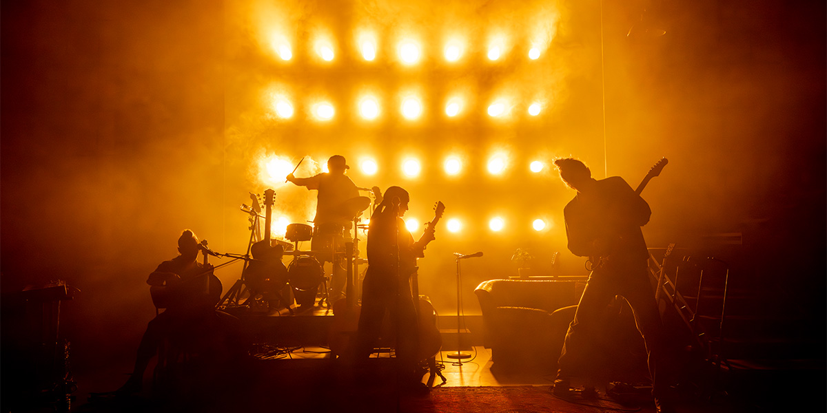 Silhouetted band members perform on stage against a backdrop of bright lights and smoke.