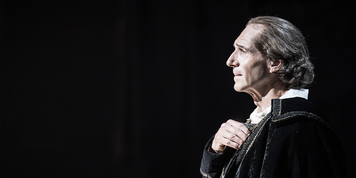 Ralph Fiennes as Henry Irving, dressed in a dark suit, gestures expressively on a dimly lit stage.