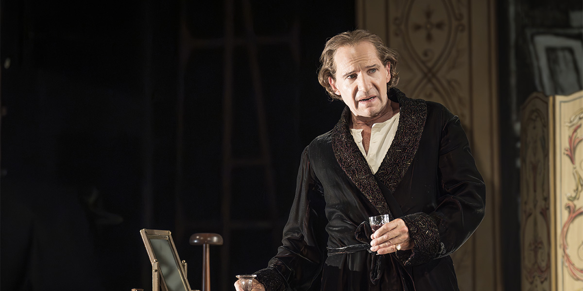 Ralph Fiennes as Henry Irving, dressed in period costume, gestures expressively on a dimly lit stage.