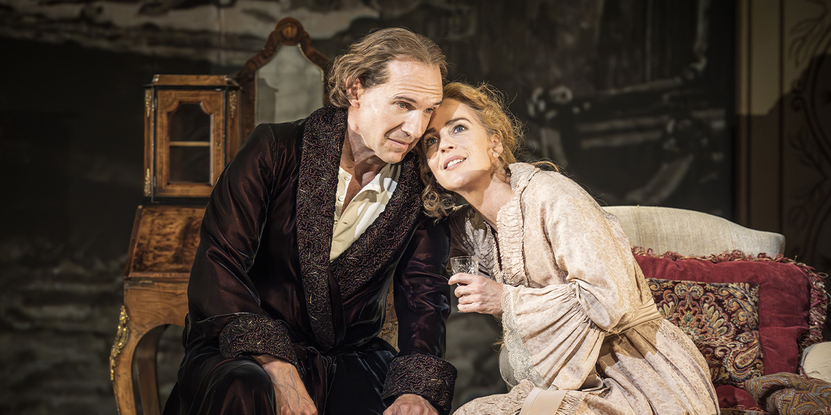 Ralph Fiennes and Miranda Raison sit together, engaged in conversation, against a softly lit, elegant backdrop.