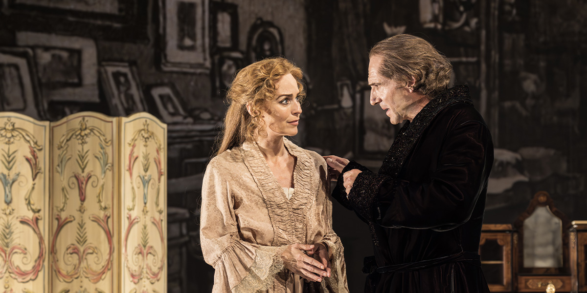 Ralph Fiennes and Miranda Raison sit together, smiling, against a soft-focus background of warm colours.