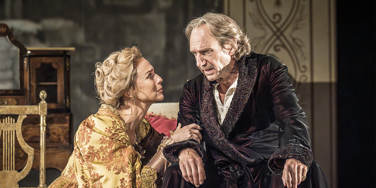 Ralph Fiennes and Miranda Raison sit together, smiling, in a warmly lit interior with wooden accents.