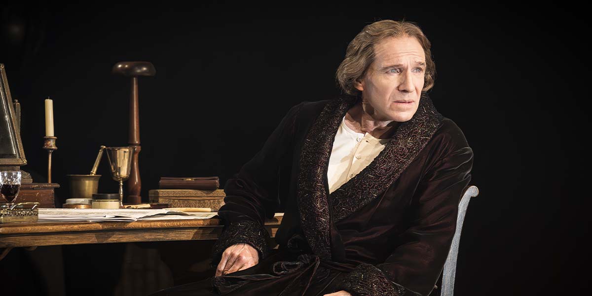 Ralph Fiennes as Henry Irving, seated at a desk, wearing a dark robe, with a contemplative expression.