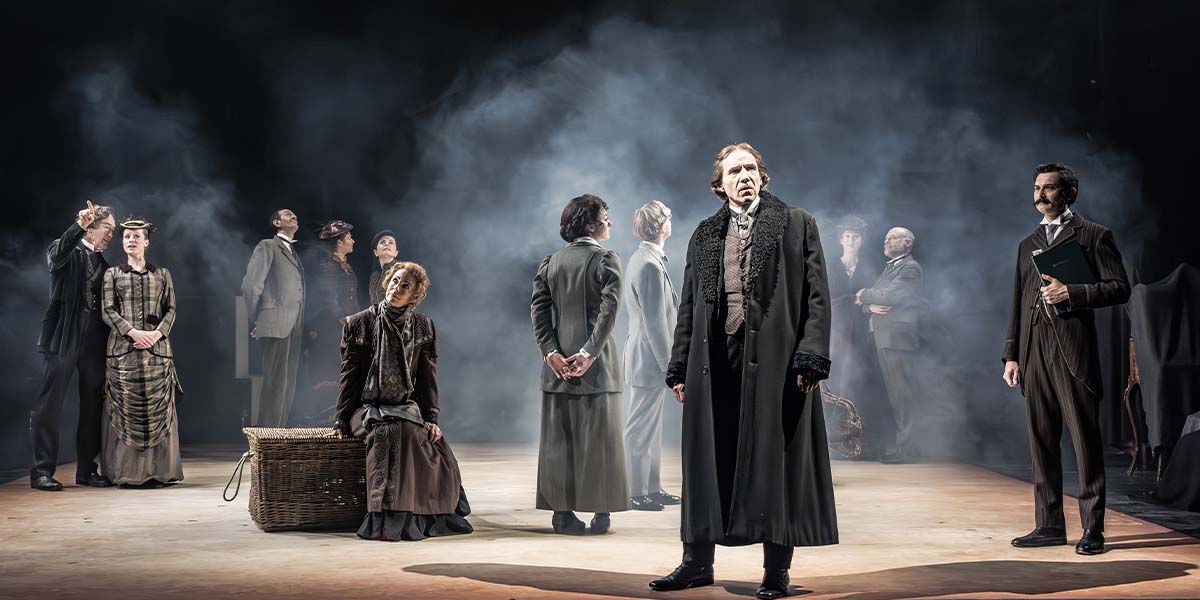 Ralph Fiennes as Henry Irving stands centre stage with an ensemble cast, dressed in period costumes.