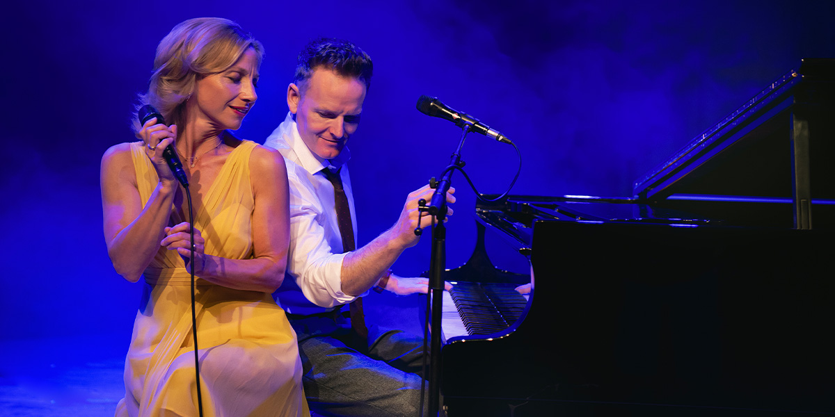 A woman in a yellow dress sings into a microphone while a man plays piano beside her.