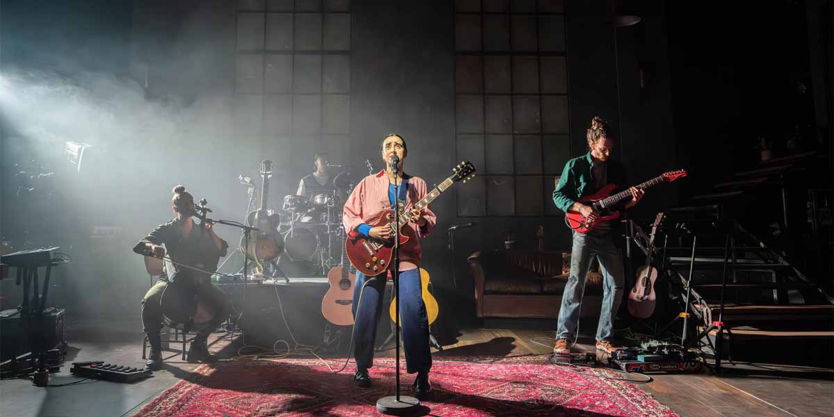 Maimuna Memon performs with the cast of Manic Street Creature, surrounded by instruments and stage lighting.