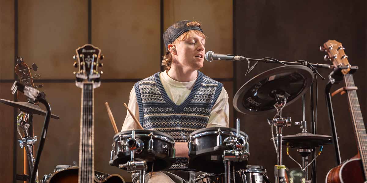 Harley Johnston, wearing a patterned vest and cap, plays drums and sings into a microphone.