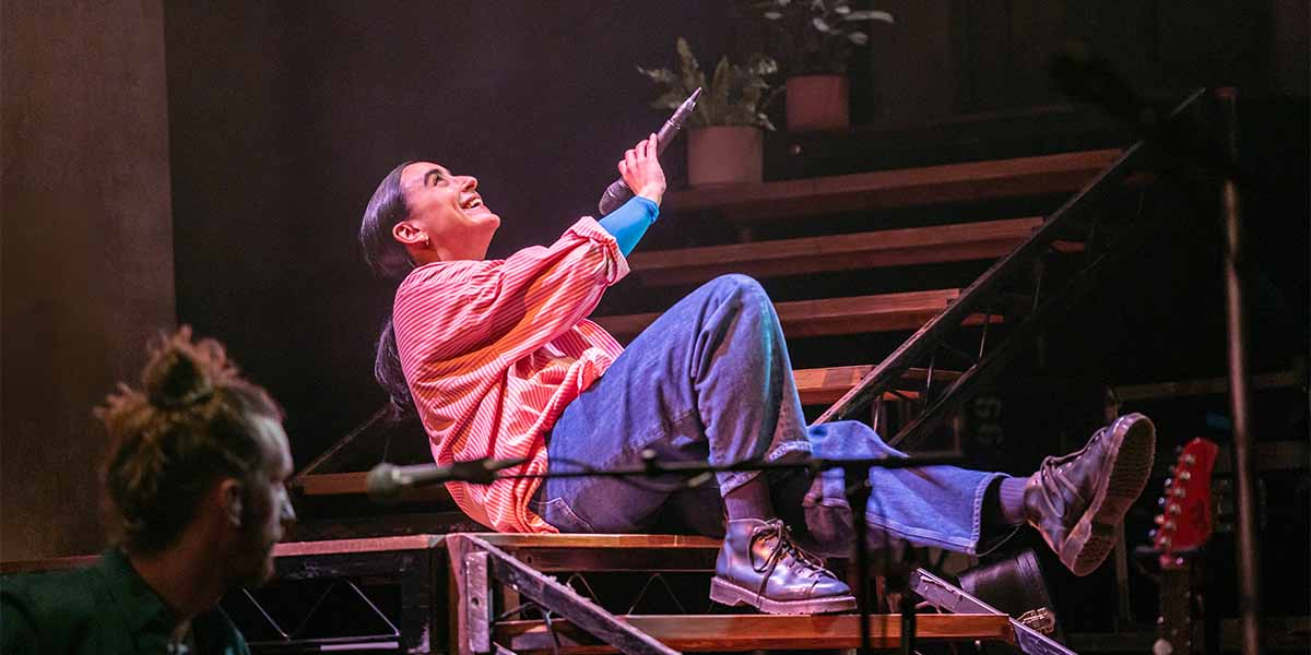 Maimuna Memon (Ria) sits on stairs, smiling and holding a microphone, wearing a striped shirt and jeans.