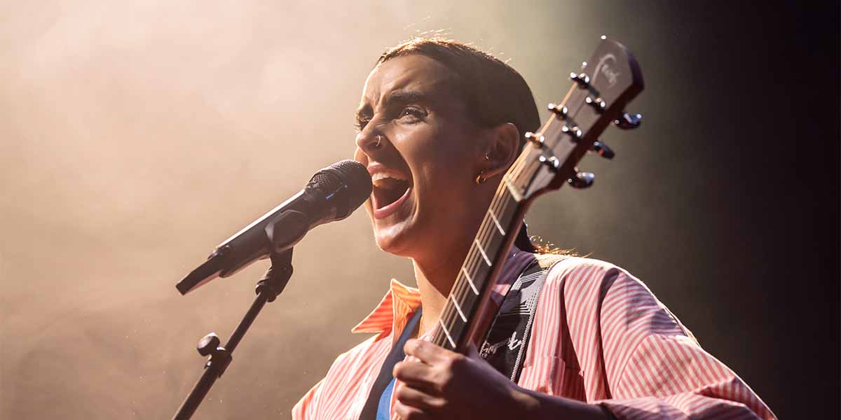 Maimuna Memon passionately sings into a microphone while holding a guitar, illuminated by soft stage lighting.