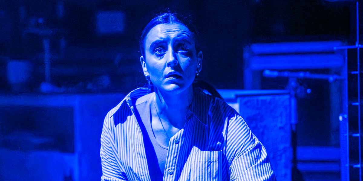 Maimuna Memon, wearing a striped shirt, gazes intensely under blue lighting in a dramatic setting.