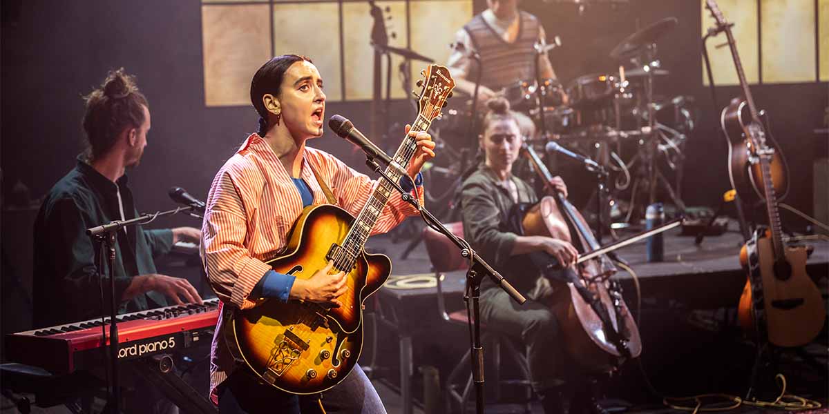 Maimuna Memon performs with a guitar, surrounded by musicians on stage, with various instruments visible.