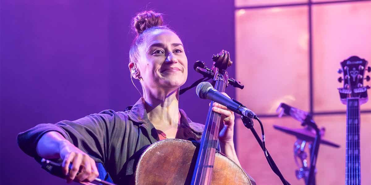 Rachel Barnes smiles while holding a cello and microphone, with purple lighting and instruments in the background.