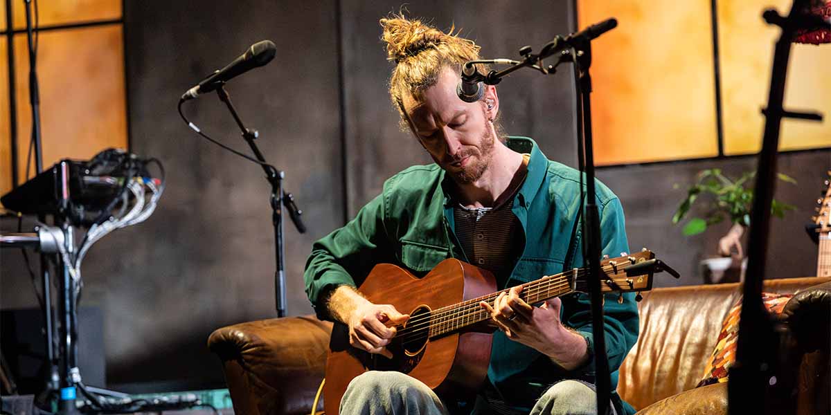 Sam Beveridge plays an acoustic guitar on a sofa, with microphones and a plant in the background.