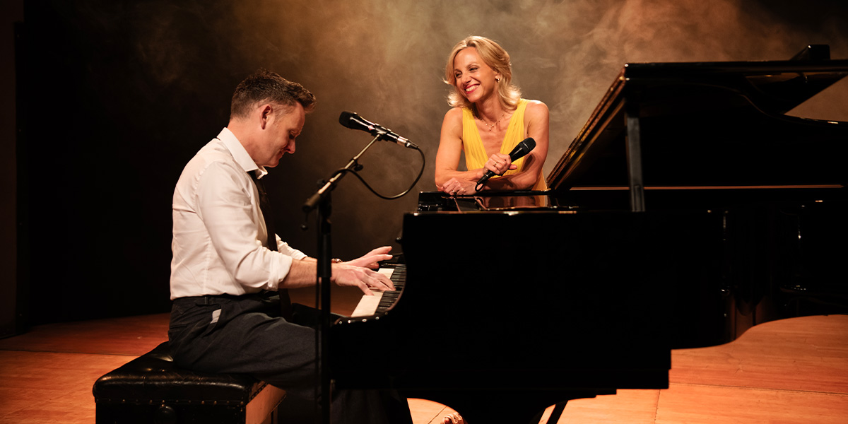 A man plays piano while a woman in a yellow dress smiles and holds a microphone.