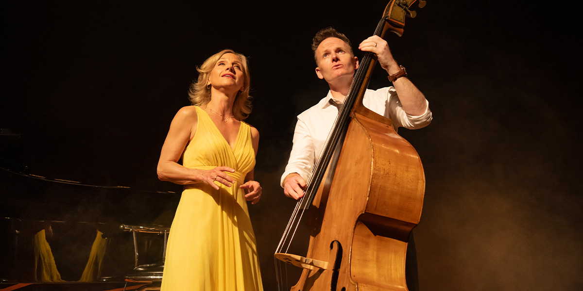 A woman in a yellow dress sings while a man plays a double bass, both illuminated by stage lights.