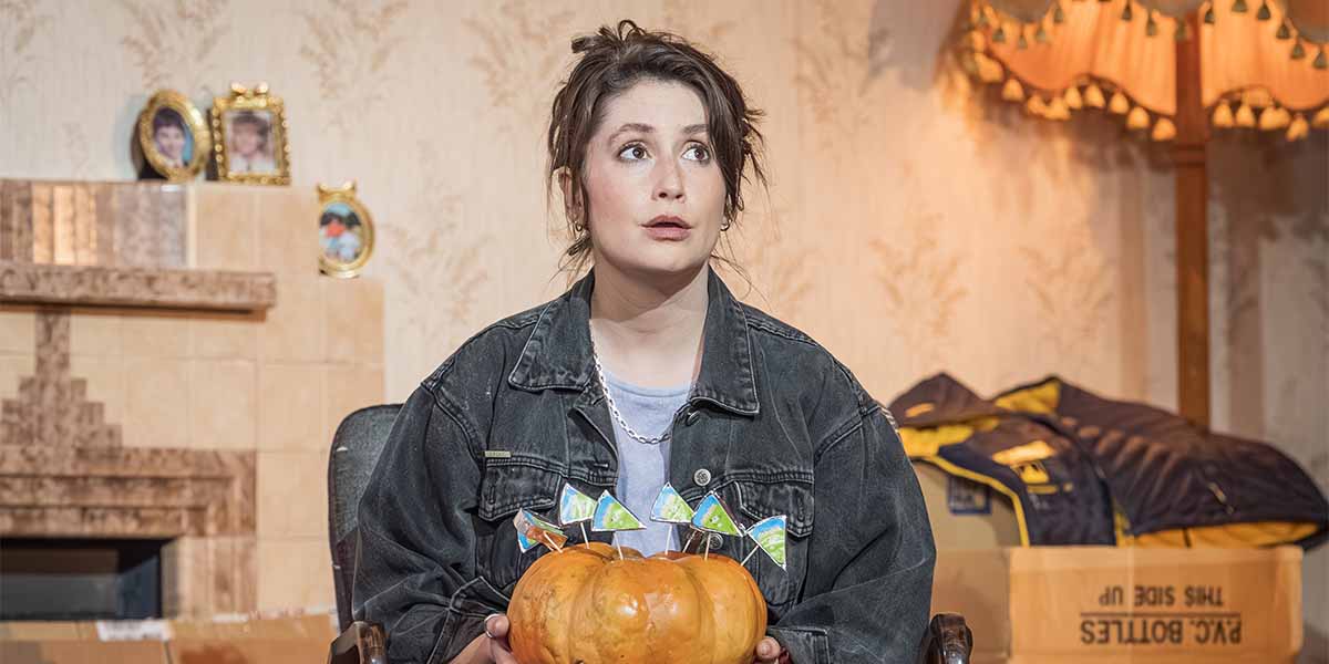 Charly Clive sits in a chair, holding a decorated pumpkin, with a patterned wallpaper background.