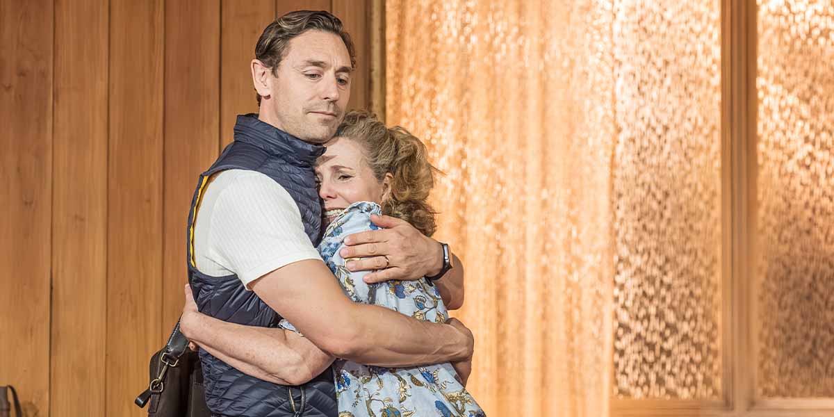 JJ Feild and Sally Phillips embrace warmly, with a wooden backdrop and soft, textured lighting.