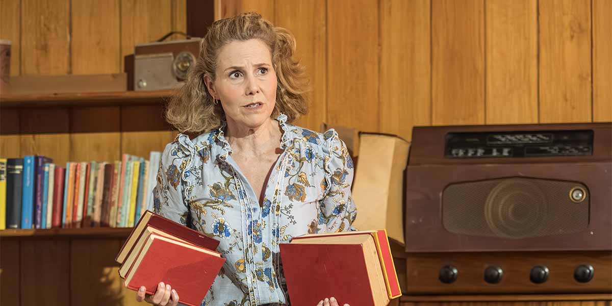Sally Phillips holds two red books, looking concerned, in a wooden room with a vintage radio.