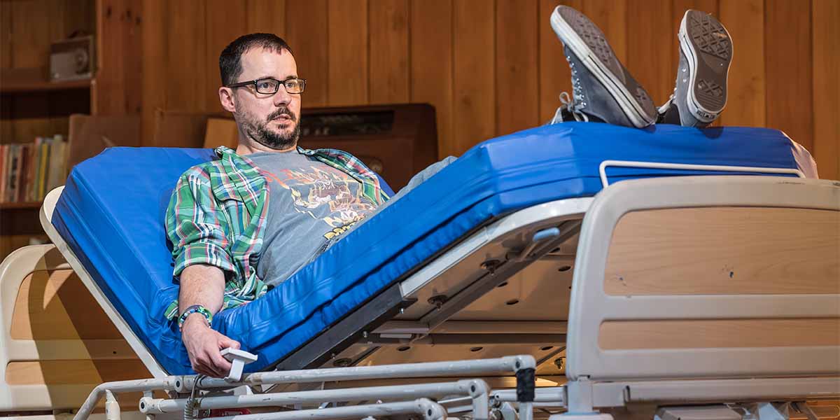 Sam Swainsbury reclines on a blue hospital bed, wearing a graphic t-shirt and glasses, with a wooden backdrop.